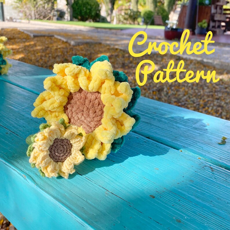 May include: Two crocheted sunflowers, one large and one small, on a blue wooden surface. The larger sunflower is yellow with a brown center and green leaves. The smaller sunflower is yellow with a brown center. The text "Crochet Pattern" is written in yellow letters on a white background.