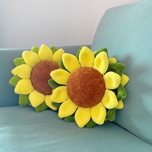 May include: Two sunflower-shaped pillows with yellow petals, brown centers, and green leaves. The pillows are made of a soft, textured material and rest on a light blue surface. The pillows are decorative.