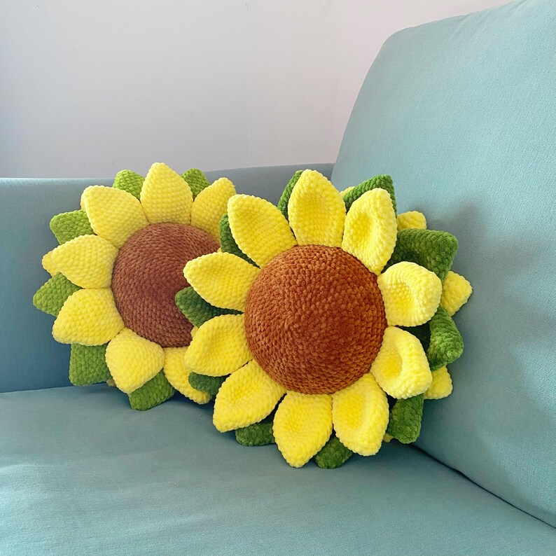 May include: Two sunflower-shaped pillows with yellow petals, brown centers, and green leaves. The pillows are made of a soft, textured material and rest on a light blue surface. The pillows are decorative.