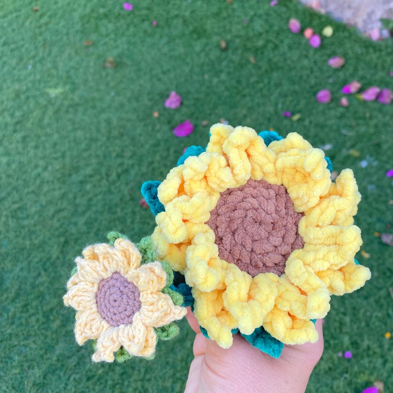 May include: Two crocheted sunflowers, one large and one small. The large sunflower is yellow with a brown center and green leaves. The small sunflower is yellow with a purple center and green leaves.