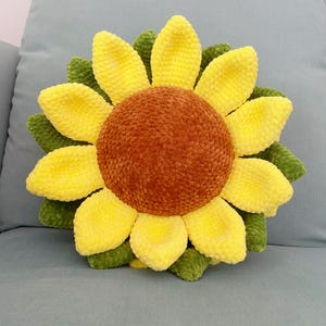 May include: A plush, sunflower-shaped pillow with a brown center and yellow petals. The petals are arranged around the center, with green leaves at the base. The pillow is designed to resemble a sunflower.