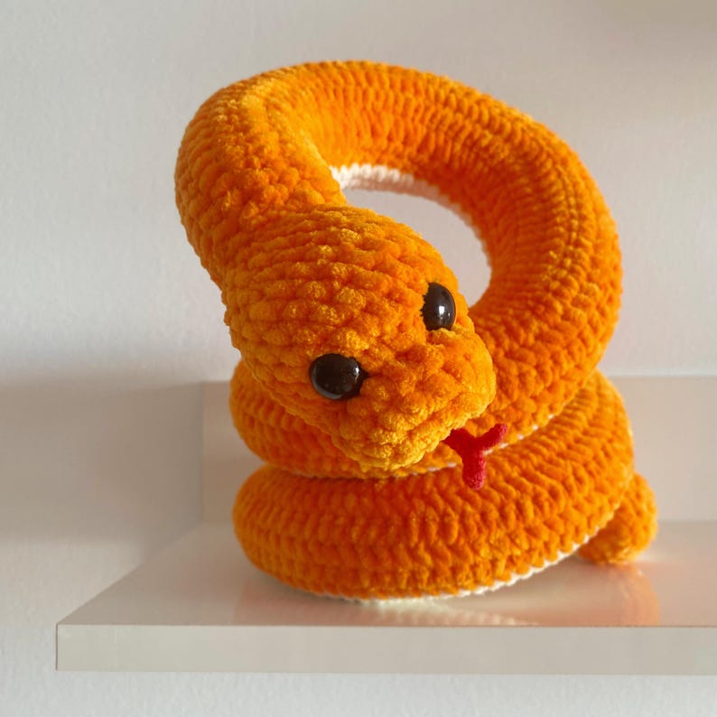 May include: A crocheted orange snake toy with black eyes and a red tongue. The snake is curled up in a circle.