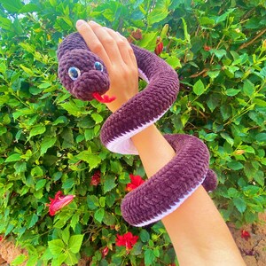 May include: A plush brown snake toy with white trim and large, black eyes. The snake is wrapped around a person's arm.