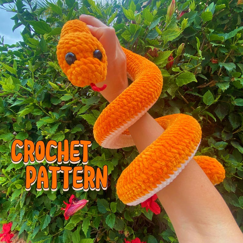 May include: Crochet pattern for a bright orange snake with white accents. The snake is curled up on a person's arm.