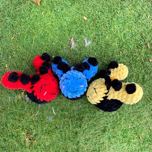 May include: Three plush, smiling caterpillar toys in red, blue, and yellow with black accents. The toys are made of a soft, textured material and are arranged on a bed of green grass. Each caterpillar has a smiling face.