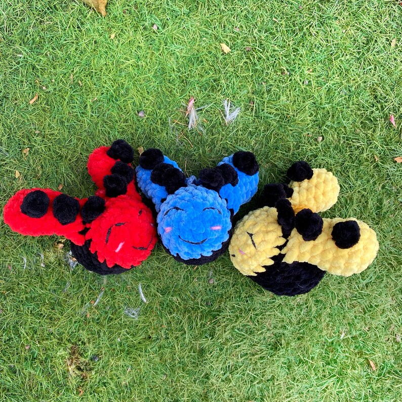 May include: Three plush, smiling caterpillar toys in red, blue, and yellow with black accents. The toys are made of a soft, textured material and are arranged on a bed of green grass. Each caterpillar has a smiling face.