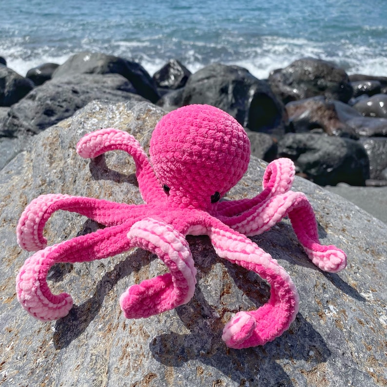 以下が含まれることがあります： 表面がテクスチャ加工されたピンクの毛糸で編まれたタコのおもちゃ。タコは灰色の岩の上に座っています。