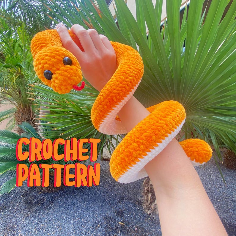 May include: A crocheted orange and white snake with black eyes. The snake is being held by a person's arm. The text "CROCHET PATTERN" is in the lower left corner of the image.