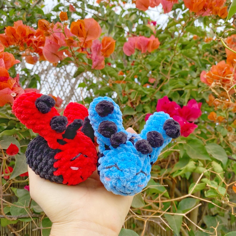 May include: Two handmade crochet creatures, one red and black, the other blue with black accents. The plush toys have large, rounded ears and are held in front of a backdrop of colorful flowers and foliage.