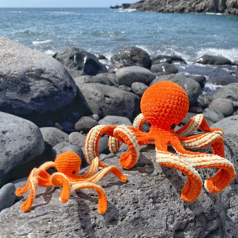 以下が含まれることがあります： 白のストライプが入ったオレンジ色の編みぐるみタコが2匹。大きいタコは8本の足、小さいタコは6本の足。タコは岩の上に座っています。