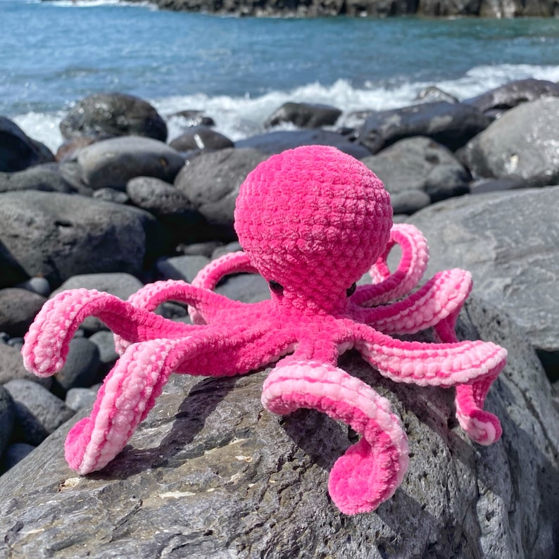 以下が含まれることがあります： 8本の触手を持つピンクの編みぐるみタコ。タコは青い海を背景に岩の上に座っています。