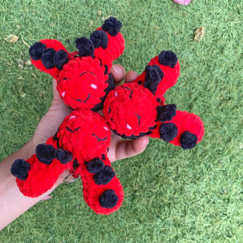 May include: Three red and black crocheted ladybug plushies. Each plushie has a round red body with black spots and wings. The ladybugs have smiling faces with white accents. The plushies are held in a hand against a green grass background.