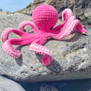 以下が含まれることがあります： 灰色の岩の上に座っている、8本の触手を持つピンクの編みぐるみタコ。