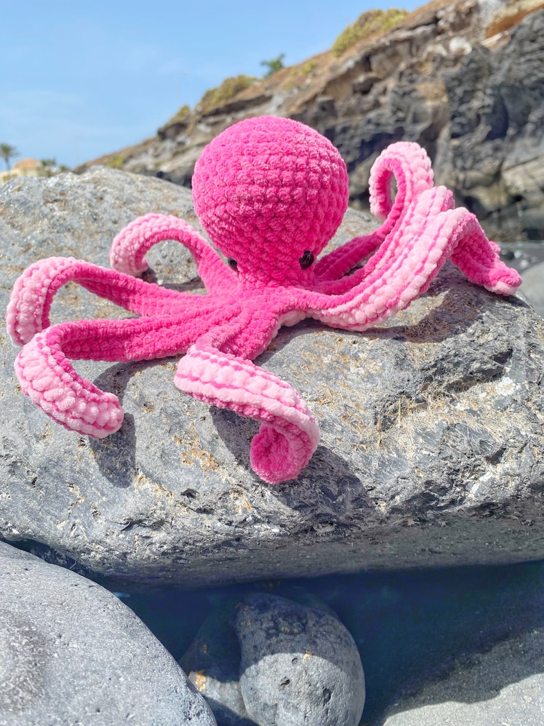 以下が含まれることがあります： 灰色の岩の上に座っている、8本の触手を持つピンクの編みぐるみタコ。