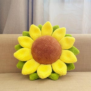 May include: A handmade sunflower-shaped pillow with a brown center, yellow petals, and green leaves. The plush pillow is designed to resemble a sunflower and is placed on a beige couch. The pillow is a decorative item.