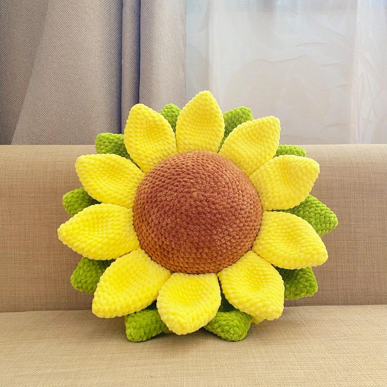May include: A handmade sunflower-shaped pillow with a brown center, yellow petals, and green leaves. The plush pillow is designed to resemble a sunflower and is placed on a beige couch. The pillow is a decorative item.