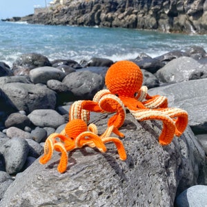 以下が含まれることがあります： 白いストライプの入ったオレンジ色の編みぐるみタコが2匹。大きなタコは岩の上に座り、触手が広がっています。小さなタコは大きなタコの前に座り、触手が丸まっています。