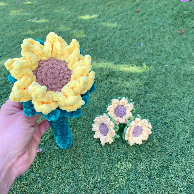 May include: A crocheted sunflower with a green stem and yellow petals. Three smaller crocheted sunflowers with yellow petals and purple centers are arranged in a cluster.