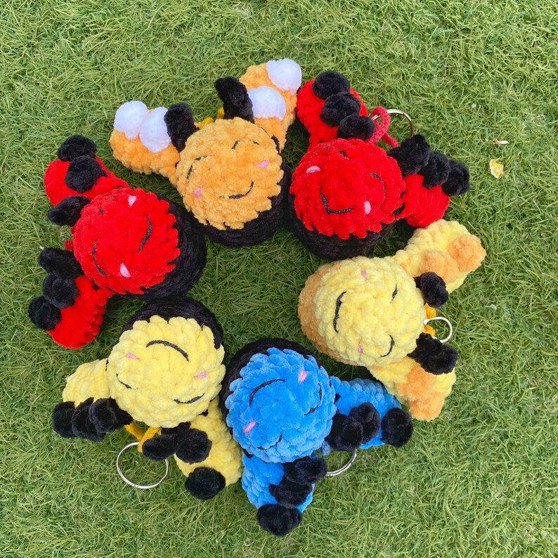May include: A collection of handmade plush bee keychains in various colors, including yellow, red, and blue. Each bee has a smiling face, black and yellow striped wings, and a silver keyring. The keychains are arranged on a bed of green grass.