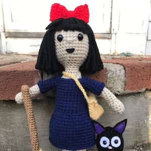 May include: A crocheted doll with black hair and a red bow, wearing a blue dress and red shoes, holding a brown broom. A black crocheted cat with white eyes sits beside the doll.