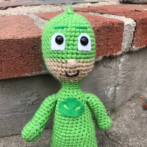 May include: Green crocheted plush toy of a superhero with a green mask and a brown beard. The toy has large black eyes and a green symbol on its chest.