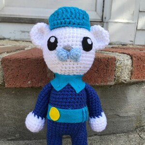 Puede incluir: Un oso de peluche blanco de crochet que lleva un gorro azul, una camisa azul y pantalones azules con un círculo amarillo en el cinturón. El oso tiene ojos negros y una nariz azul.