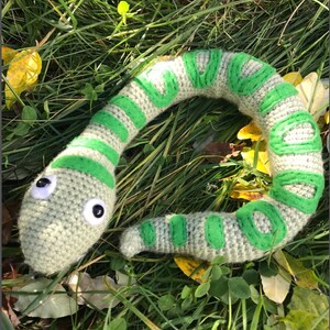 Könnte beinhalten: Ein grünes und graues Häkel-Schlangenspielzeug mit großen schwarzen Augen. Die Schlange liegt auf einem Bett aus grünem Gras.