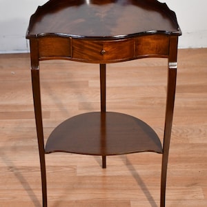 May include: A dark brown wooden side table with a semi-circular top featuring a scalloped edge, a single central drawer with a small round knob, and a lower semi-circular shelf supported by three slender legs with a polished wood grain finish.