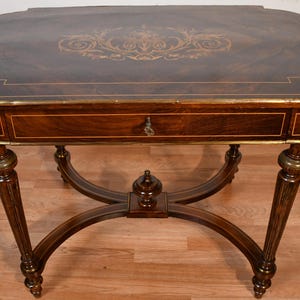 May include: A dark brown antique table with a decorative inlaid top and a single drawer. The table has ornate legs and brass accents. The top features a gold-colored floral design. The table is approximately 30 inches long.
