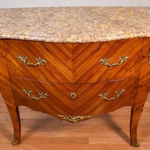May include: A wooden chest of drawers with a marble top. The chest has a curved front with two drawers and ornate brass handles and accents. The wood grain is a chevron pattern in shades of brown. The marble top has a mottled pattern of beige and brown.