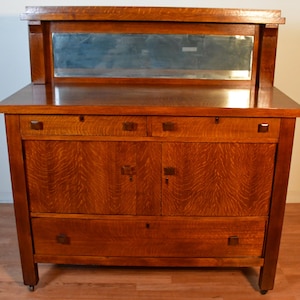May include: A wooden dresser with a mirror, featuring a warm, reddish-brown finish. The dresser has two drawers, two cabinet doors, and a lower drawer. The mirror is rectangular and set above the dresser top. The wood grain is visible.