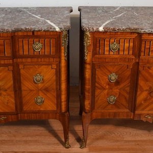 May include: Two antique wooden cabinets with marble tops. The cabinets have a warm brown finish with intricate inlaid patterns and brass hardware. Each cabinet features drawers and doors, with decorative floral motifs on the front.