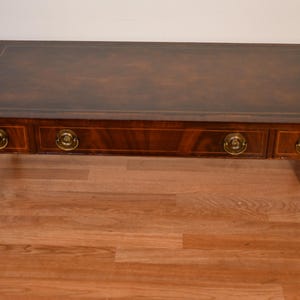 May include: A rectangular wooden coffee table with a dark brown finish and a leather-like top inlay. The table features two drop-leaf sides, four brass ring pulls on the front, and tapered legs with light-colored wood inlay detailing.