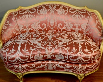 May include: A vintage loveseat with a curved back and ornate cream-colored frame. The seat and back are upholstered in a reddish-brown fabric with a repeating silver floral and mythical creature pattern. The sofa has a classic, elegant design.