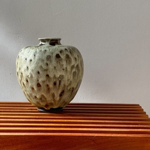 以下が含まれることがあります： 斑点のある釉薬をかけた緑と茶色の陶器の花瓶。花瓶は丸みを帯びた形をしていて、上部に小さな開口部があります。