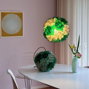 May include: A modern dining room scene features a white table, a wire chair, and a painting with a yellow frame. Two decorative floral lamps, one hanging and one on the table, provide soft lighting. A vase with flowers adds a touch of nature.