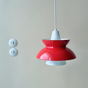 May include: A red and white pendant light fixture with a unique, layered design. The light has a glossy red shade and a white base and top. Two white light switches are mounted on the wall to the left.