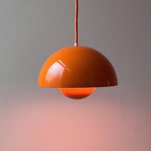 以下が含まれることがあります： 光沢のあるオレンジ色のペンダントライト。大きな半球状のシェードと、その下に吊り下げられた小さな同色のオレンジ色のドームで構成されています。オレンジ色の布製コードで吊り下げられており、中心部から温かく柔らかな光を放っています。