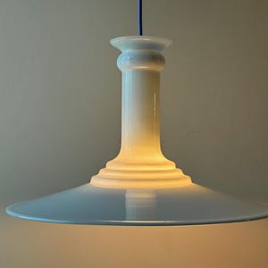 May include: A white pendant light with a wide, flat, circular shade and a tall, cylindrical base. The light is turned on and emits a warm, yellow glow.
