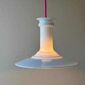 Peut inclure: Une suspension en céramique blanche dotée d'un large abat-jour évasé et d'un col cylindrique. La lampe est suspendue par un cordon en tissu rose vif. La surface de la céramique est lisse et brillante, reflétant la lumière avec un éclat doux.