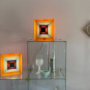 May include: Two square-shaped lamps with layered orange, white, blue, and red glass panels. The lamps are on a glass shelf with other decorative objects.
