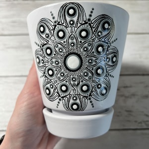 May include: A white ceramic plant pot with a detailed black and gray mandala design. The pot has a matching saucer and is held by a hand. The mandala pattern features concentric circles and dot patterns, creating a symmetrical floral design.