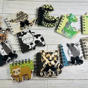 May include: A collection of animal-themed spiral notebooks. Designs include a giraffe, alligator, cow, snake, zebra, sloth, leopard, and a second zebra and alligator. Each notebook has a unique animal design and a black bow.