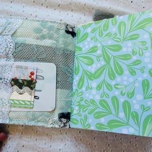 K&ouml;nnte beinhalten: Ein kleines, handgemachtes Journal mit einem blauen und gr&uuml;nen Blumenmuster auf dem Einband. Das Journal hat einen wei&szlig;en Spitzenbesatz und eine transparente Plastik-Tasche auf dem vorderen Einband. Die Tasche enth&auml;lt ein kleines St&uuml;ck gemustertes Papier mit roten Beeren.
