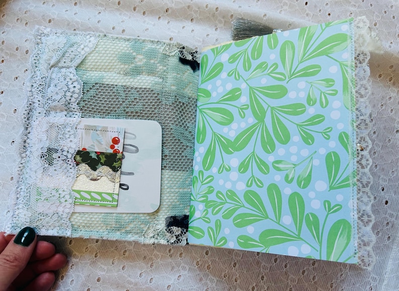 K&ouml;nnte beinhalten: Ein kleines, handgemachtes Journal mit einem blauen und gr&uuml;nen Blumenmuster auf dem Einband. Das Journal hat einen wei&szlig;en Spitzenbesatz und eine transparente Plastik-Tasche auf dem vorderen Einband. Die Tasche enth&auml;lt ein kleines St&uuml;ck gemustertes Papier mit roten Beeren.