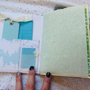 K&ouml;nnte beinhalten: Ein hellblaues und wei&szlig;es Journal mit dekorativem Papier und Spitzenakzenten. Das Journal hat einen hellgr&uuml;nen Papiereinsatz und einen kleinen Metallglockenanh&auml;nger.