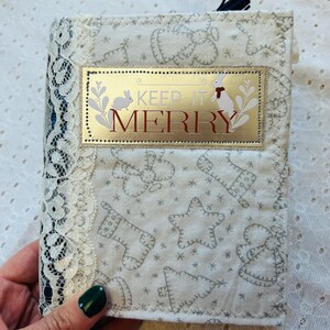 K&ouml;nnte beinhalten: Ein wei&szlig;es Stoffjournal mit einem goldenen und roten Etikett, auf dem "Keep it Merry" steht. Das Journal hat einen wei&szlig;en Spitzenbesatz und ein schwarzes Band.