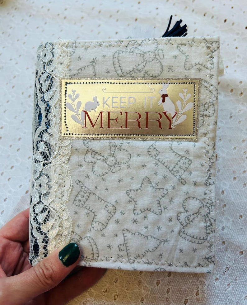 K&ouml;nnte beinhalten: Ein wei&szlig;es Stoffjournal mit einem goldenen und roten Etikett, auf dem "Keep it Merry" steht. Das Journal hat einen wei&szlig;en Spitzenbesatz und ein schwarzes Band.
