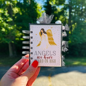 May include: A small, spiral-bound notebook with a cover featuring an angel illustration with gold wings. The text "ANGELS we have HEARD ON HIGH" is printed below the angel. The notebook has a black border and decorative silver ribbon.