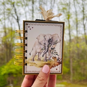 May include: A small, spiral-bound notebook with a gold-colored metal binding. The cover features a watercolor illustration of two elephants with floral accents. The notebook is held in front of a blurred green background.
